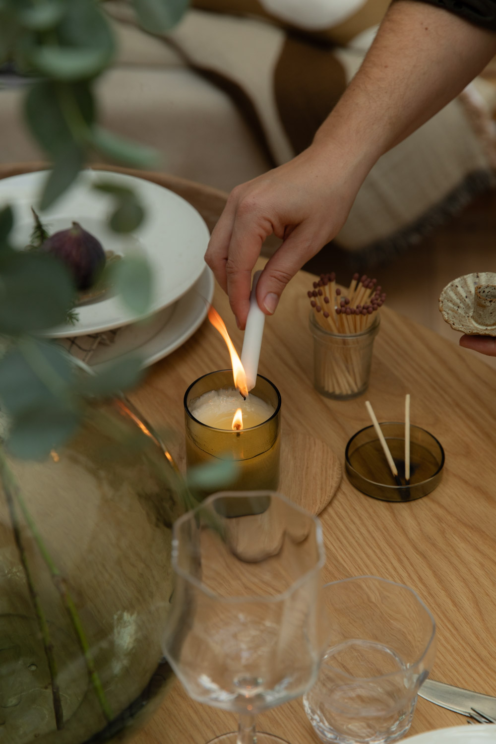 A hand lights a scented candle, set on a wooden table with other objects.